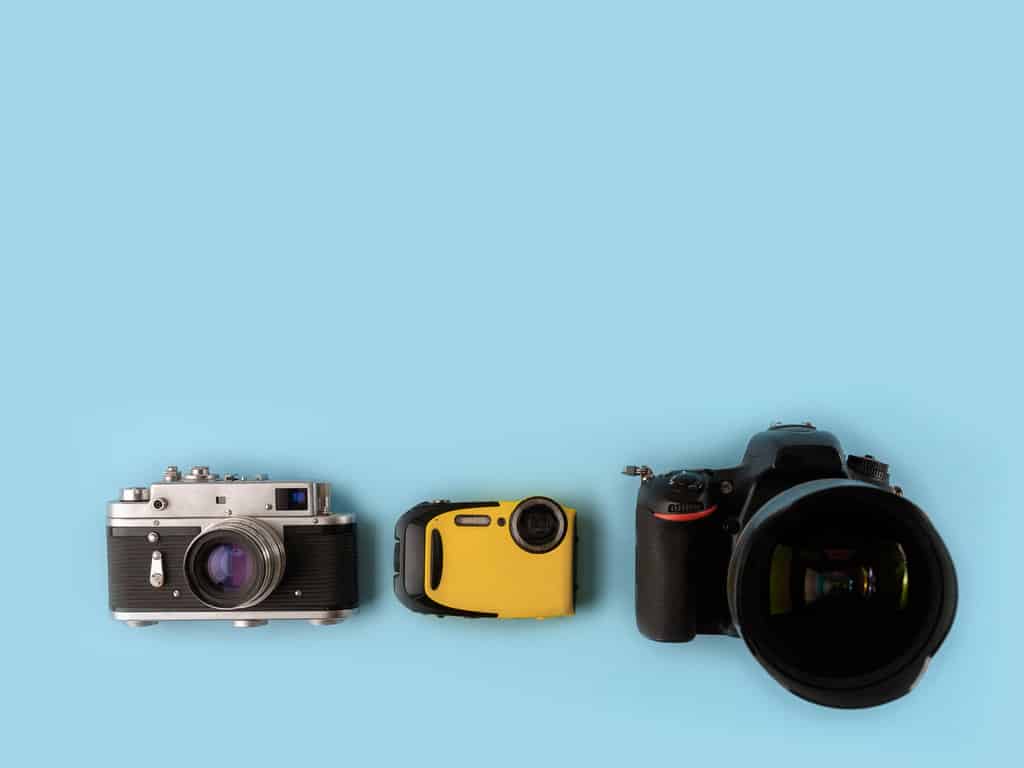 sur un fond bleu ciel, 3 appareils photos de générations différentes sont alignés. Un argentique à gauche, un appareil jetable au milieu et un réflexe à droite.