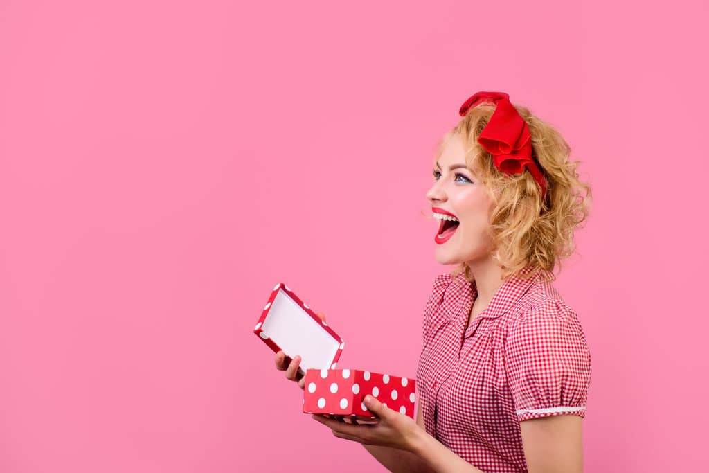 une pinup est de profil sur un fond rose. la bouche ouverte de joie, elle ouvre une boite rouge à pois blancs. elle porte un headband rouge dans ses cheveux et un chemisier à carreaux rouge et blanc.