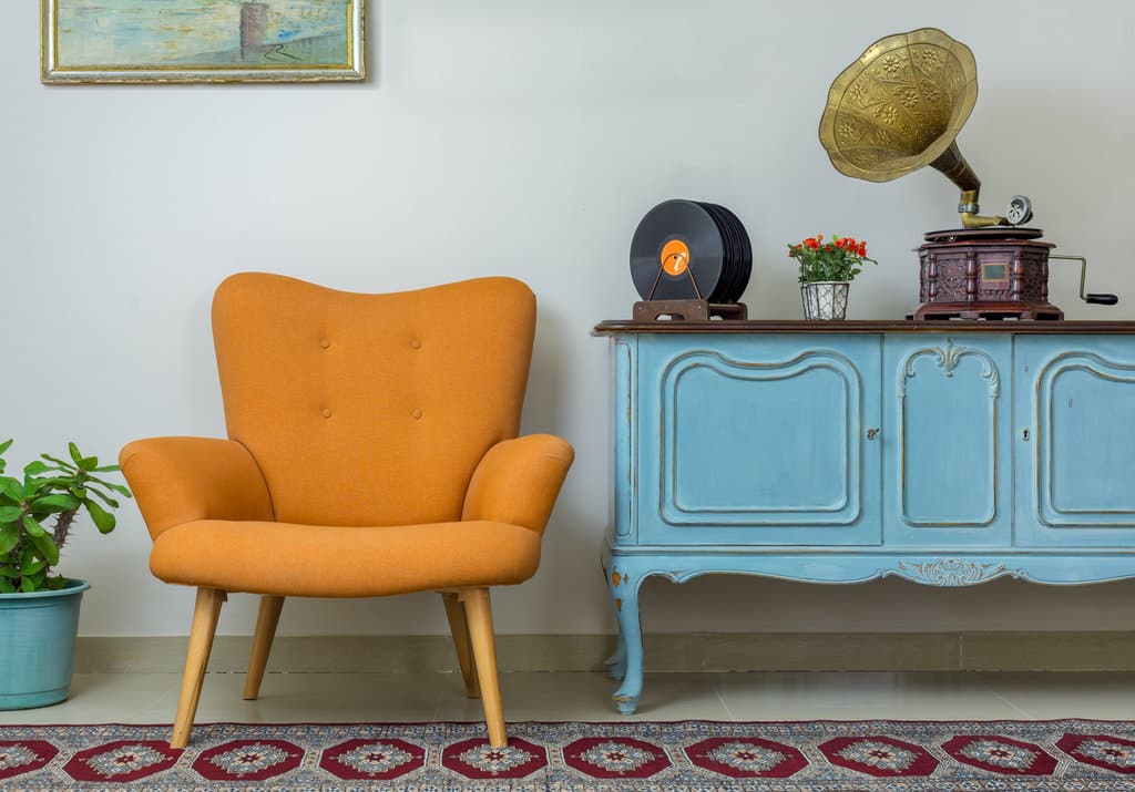 vue sur des meubles vintage dont un fauteuil orange et une commode en bois peinte en bleue. un vieux tourne disque et posé sur la commode.