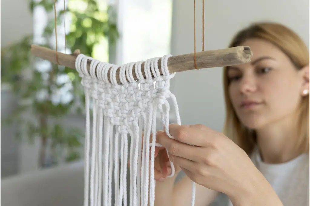 une jeune femme confectionne des noeuds de macramé à l'aide de fils épais blanc accroché à un bout de bois suspendu en l'air par des fils fins