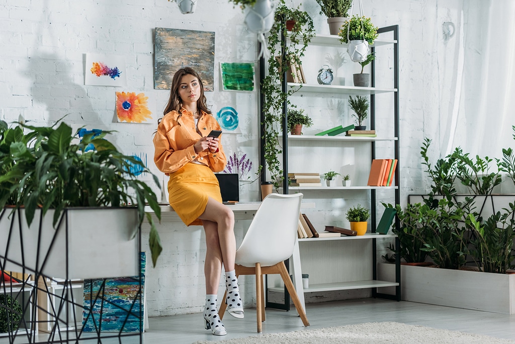 une jeune femme en robe vintage orange est assise sur un bureau en tenant un téléphone dans se mains. des plantes vertes sont posées un peu partout dans la pièce et notamment sur une étagère.