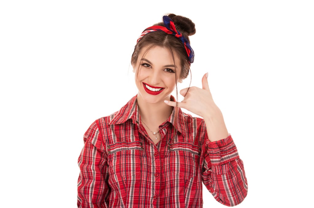 une jeune femme sourit et fait le signe du téléphone avec sa main. elle porte un chemisier à rayures rouge et un foulard rouge et bleu.
