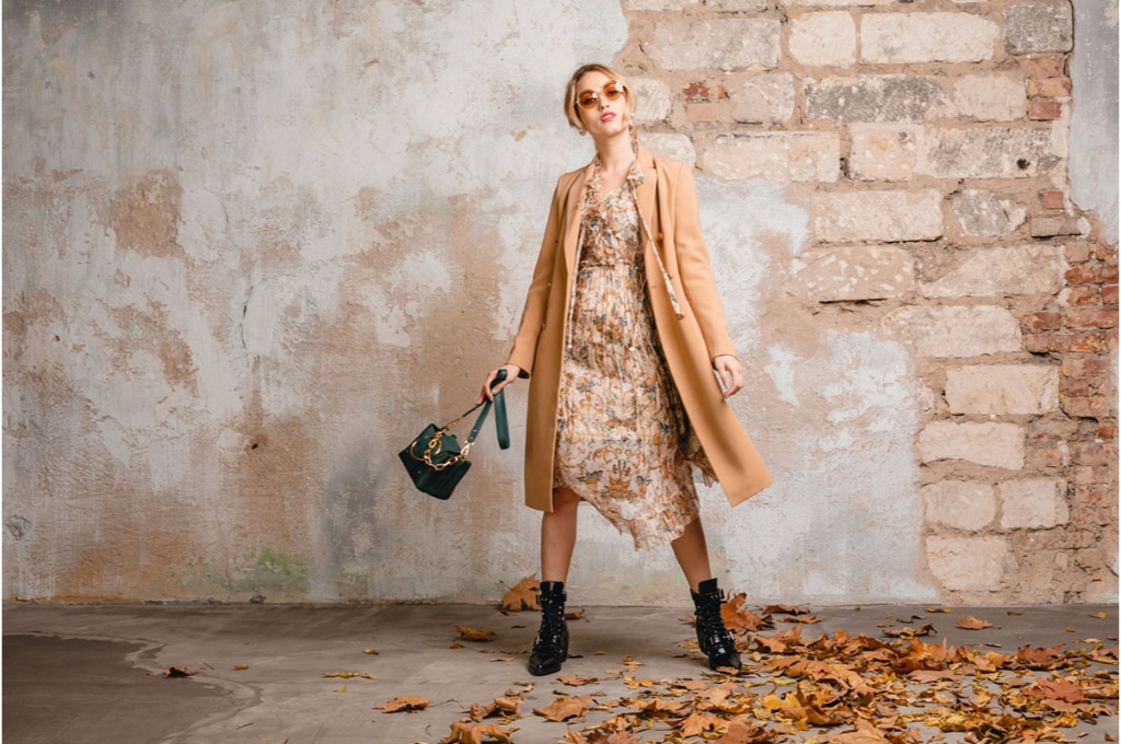 une femme en robe longue à motifs et manteau long beige est devant un mur de briques. elle porte des chaussures noires et des lunettes marron vintage.