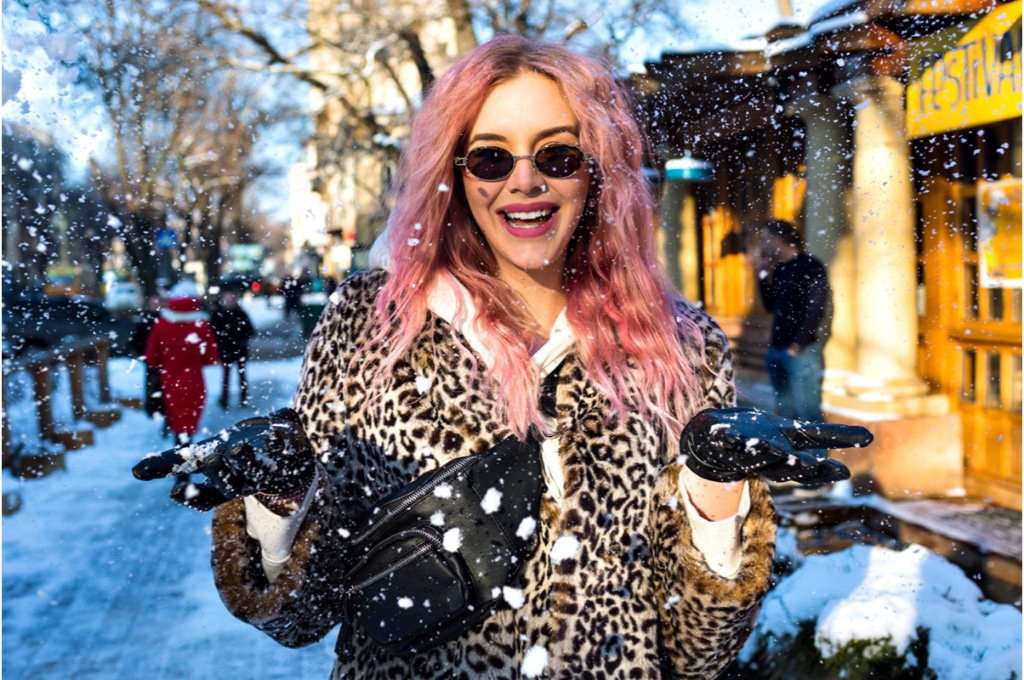 une femme aux cheveux gauffrés roses sourit sous la neige qui tombe. elle porte un manteau léopard et des gants noirs. une banane noire est accroché devant sa poitrine. elle porte des lunettes de soleil vintage.
