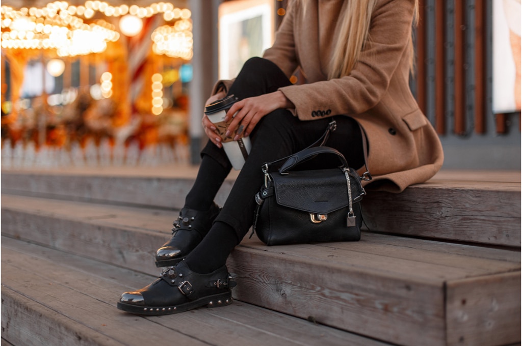une femme en manteau beige et legging noir est assise dehors sur des marches en bois. elle tient un gobelet de boisson chaude entre ses mains. son petit sac en cuir noir est posé près d'elle.