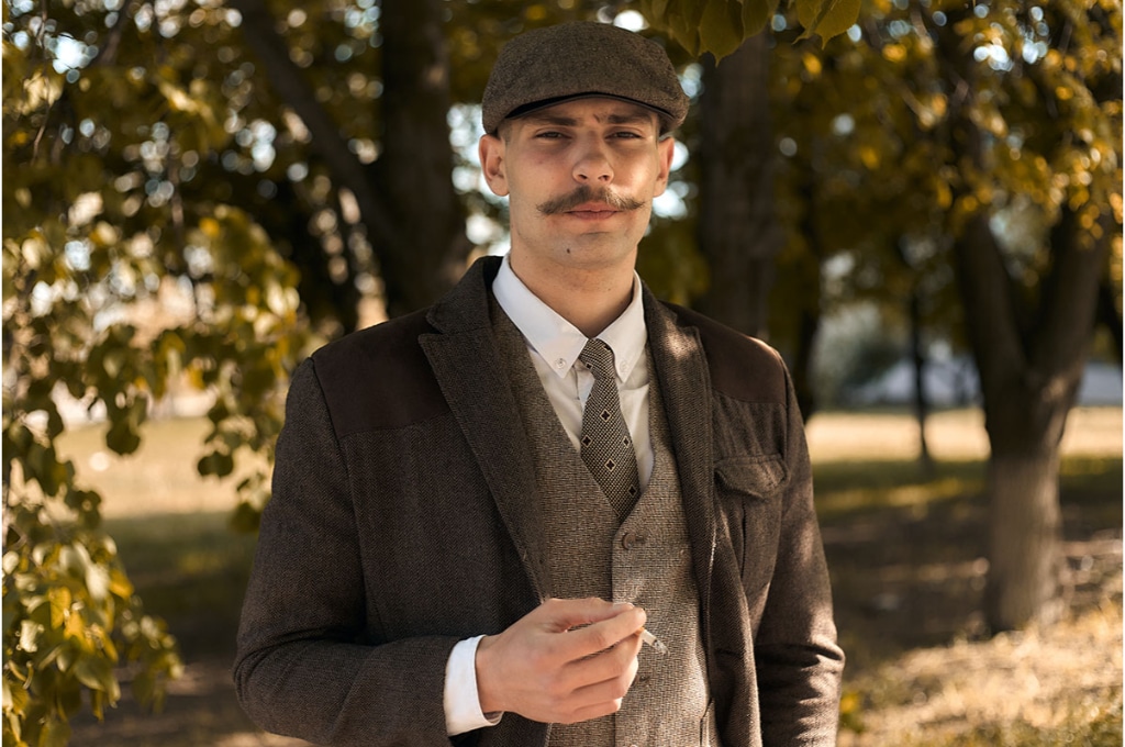 un homme en costume marron avec une moustache, porte une casquette irlandaise. il tient une cigarette allumée dans sa main droite.