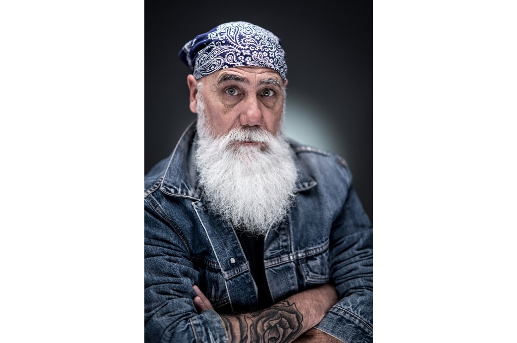 un homme avec une barbe blanche croise les bras et regarde le photographe. il porte une veste en jean et un bandana bleu sur la tête. il a un tatouage d'une rose sur son bras droit.