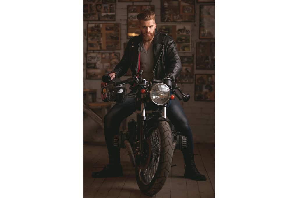 un motard est assis sur sa moto vintage face au photographe. il porte une veste en cuir et a les cheveux longs avec une barbe.