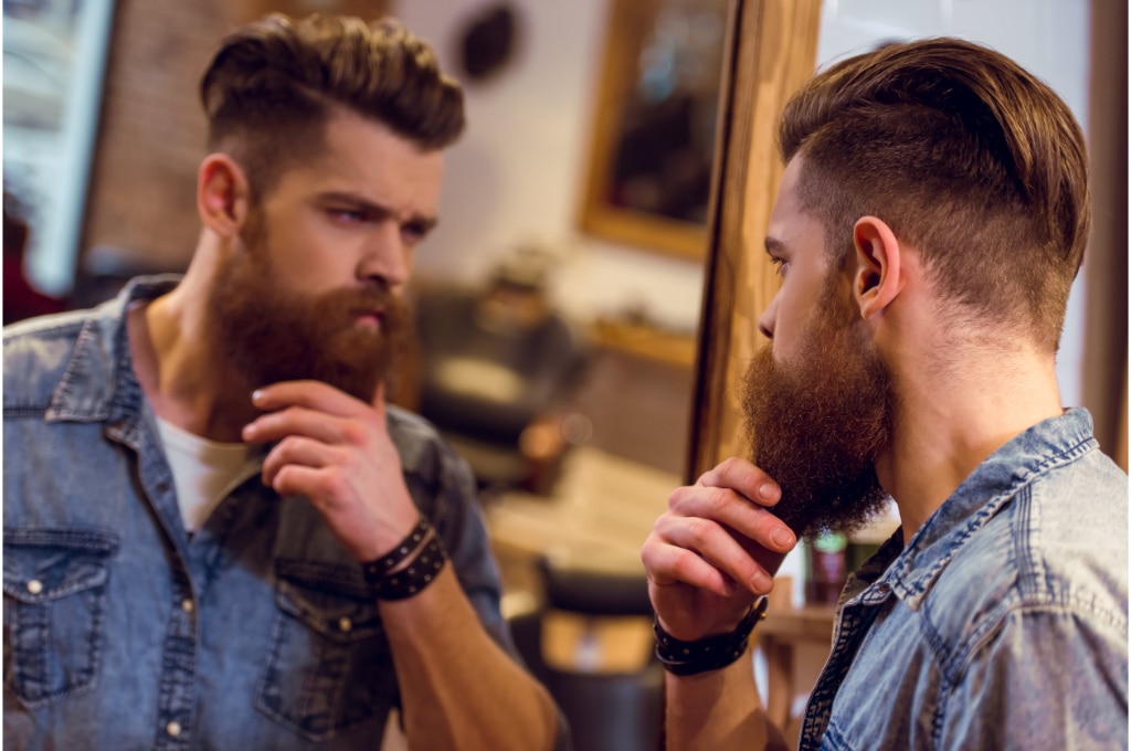un homme se regarde et se touche la barbe devant un miroir. ses cheveux sont longs au-dessus de la tête et courts sur les côtés. il porte une chemise en jean.