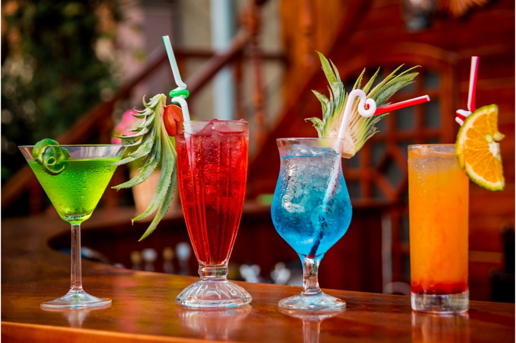 4 cocktails colorés sont posés sur une table de bar en bois. le verre de gauche est vert avec une tranche de citron vert dessus, le deuxième est rouge avec une paille entortillée et un bout d'un fruit. Le troisième verre est bleu avec une paille entortillée et un fruit. Le dernier verre est orange avec une paille entortillée et un morceau d'orange dessus.