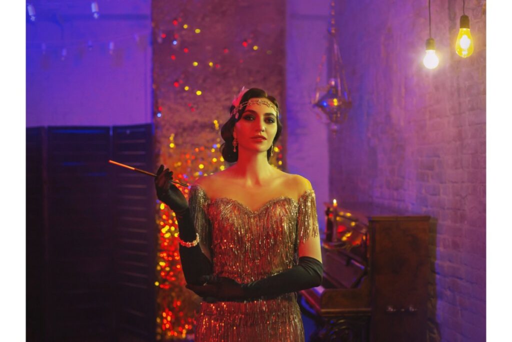 une femme vêtue et maquillée comme dans les années 20 porte une robe brillante avec de longs gants noirs. Elle tient un porte-cigarette à la main.