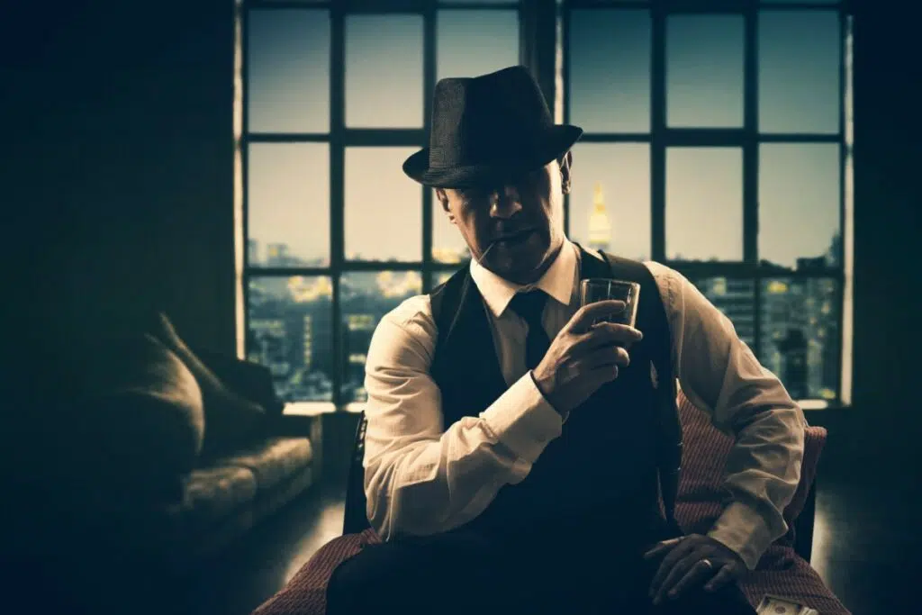 un homme en tenue charleston tient un verre à la main. il est assis dans un fauteuil dans un salon. il porte un chapeau noir, une chemise blanche avec une cravate et un veste sans manche.