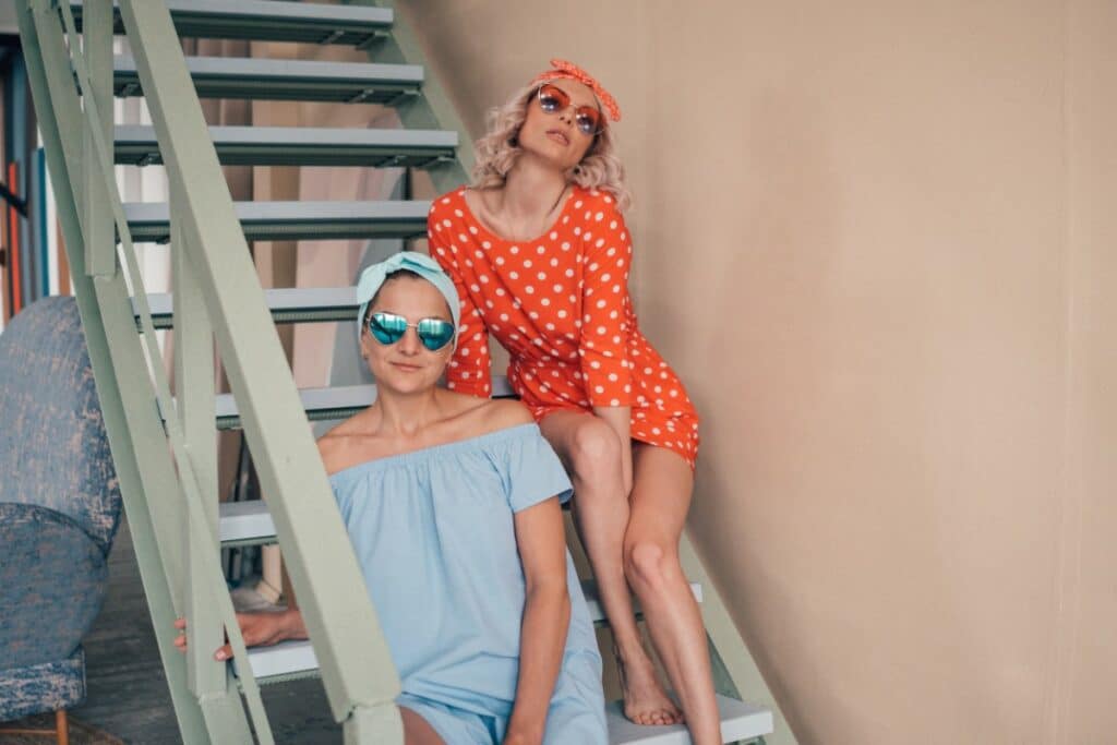 2 femmes prennent la pose assises sur des marches d'escalier. une porte une robe orange à pois blanc, un bandeau noué dans les cheveux et des lunettes oranges. l'autre est en robe bleue pastel avec les épaules découvertes, un bandeau bleu dans les cheveux et des lunettes bleues.