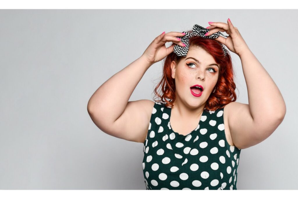 Quelle robe choisir pour une soirée guinguette ? Mode et beauté une femme ronde en robe noire à pois blancs noue un bandeau noir et blanc dans ses cheveux roux bouclés. elle a une bouche maquillée ouverte. elle porte du vernis à ongle rose.