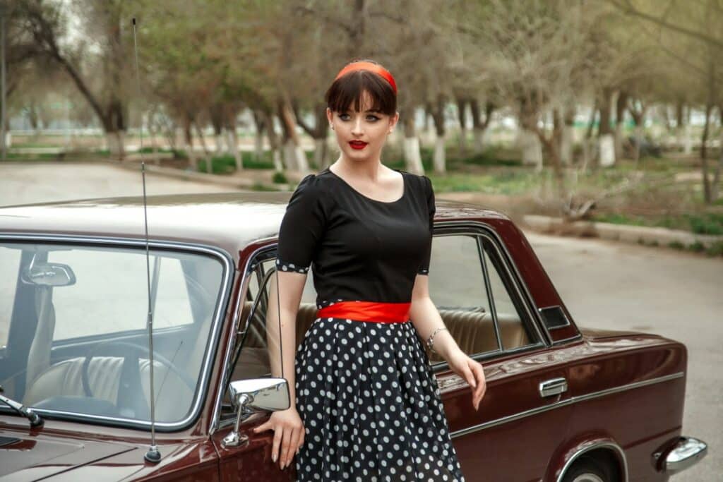 appuyée sur une voiture vintage, une femme est habillée rétro. elle porte une robe avec le haut noir et le bas de la robe noir à pois blancs. elle porte une ceinture rouge et un bandeau rouge dans les cheveux.