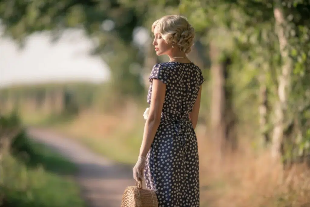 une femme blonde en tenue rétro est de dos dehors sur un chemin. elle porte un sac en osier et une robe à pois.