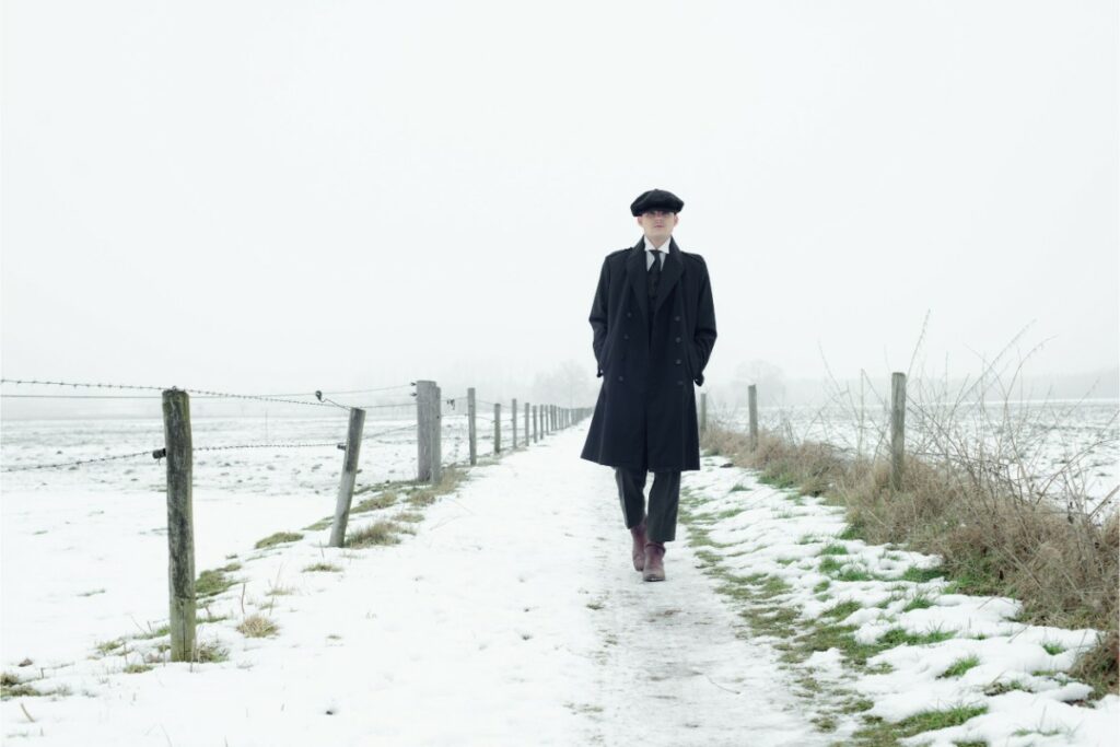 dans un paysage de campagne enneigée, un homme habillé en style rétro 1920 avec un long manteau noir et une casquette gavroche.
