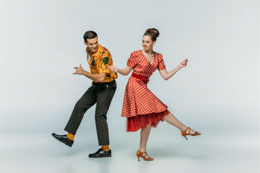 un homme et une femme dans le style rockabilly, dansent ensemble le rock sur un fond blanc. la femme porte une robe rouge à pois blancs et une ceinture rouge. l'homme porte une chemise jaune à motifs et un pantalon gris foncés.