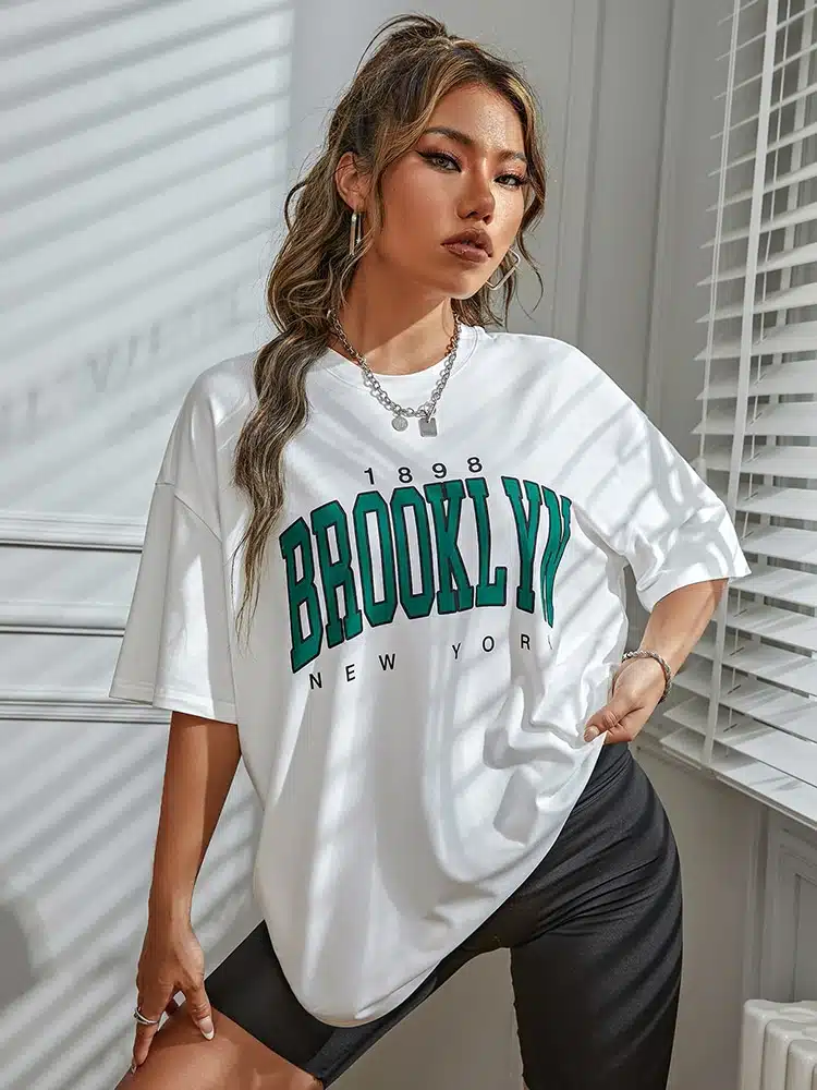 T-shirt Vintage de couleur blanc avec l'inscription Brooklyn en vert porté par une femme devant un mur blanc
