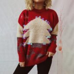 Femme blonde debout devant un mur gris, portant un jean noir et un pull de Noël rouge avec un imprimé sapin de Noël en relief et des paillettes