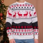 Homme debout de face, devant un sapin dans un salon, portant un jean et un pull blanc de Noël à motifs